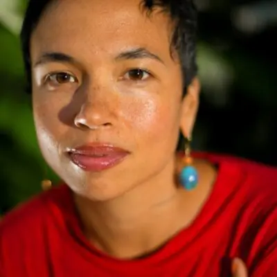 Color portrait photo of Holland Andrews. They are against a green background, wearing a red top and blue earrings.