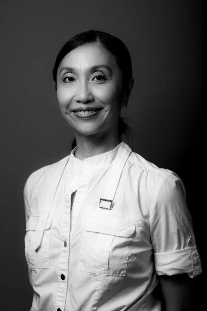 Black and white portrait photo of Janet Wong. They are smiling at the camera with their arms behind their back, wearing a white collarless button up shirt.