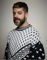Color portrait photo of Miguel Gutierrez. They are facing the camera, wearing a black and white sweater with a geometric knit pattern against an off-white background.