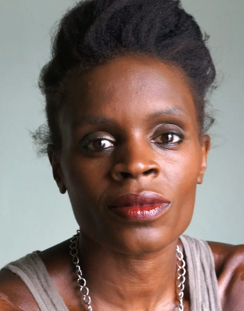 Color portrait photo of Okwui Okpokwasili. They face the camera against a grey background, wearing a grey tank top and silver necklace.