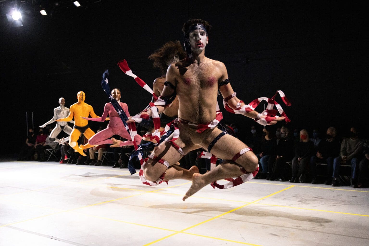 A line of dancers performs on a floor level white stage with the audience in the background. The performers are wearing body paint, minimal clothing, with the closest dancer wearing American flag ribbons on their body. The dancers are in a dynamic pose, caught jumping in the air with their legs tucked underneath themselves.