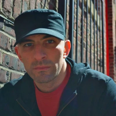 Color headshot of Adrian Silver. They are facing the camera, outside by a brick wall, wearing a black hat and swetshirt.