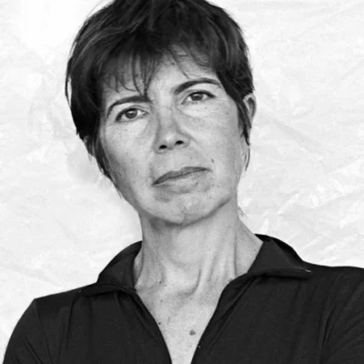 Black and white photo portrait of Elizabeth Diller. They are standing against a white stucco wall wearing a black collared shirt.