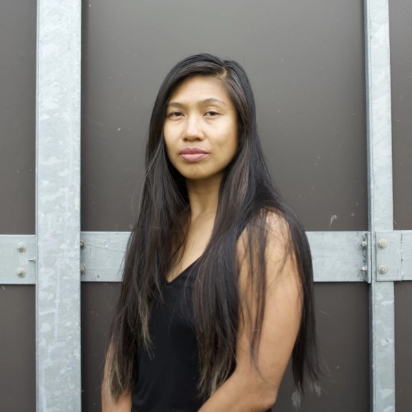 Headshot photo of Kristel Baldoz. They are sitting portrait style against a brown wall with metal beams, facing the camera. They are wearing a black dress.
