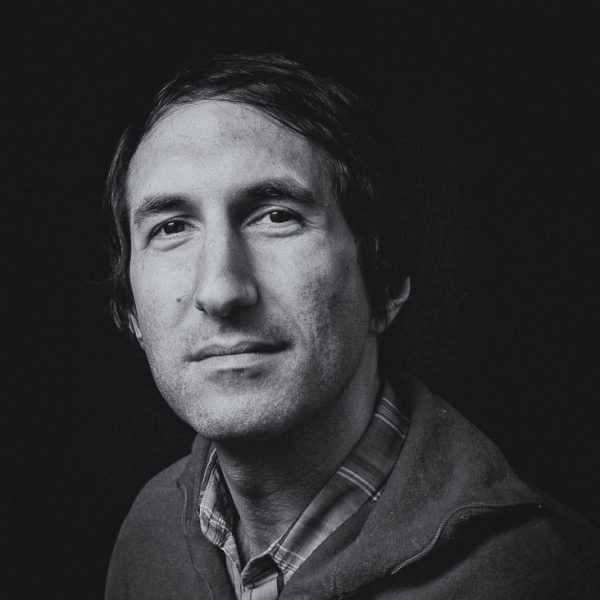 Black and white photo portrait of Nick Hallet. They face the camera with a flannel shirt and a black sweater.