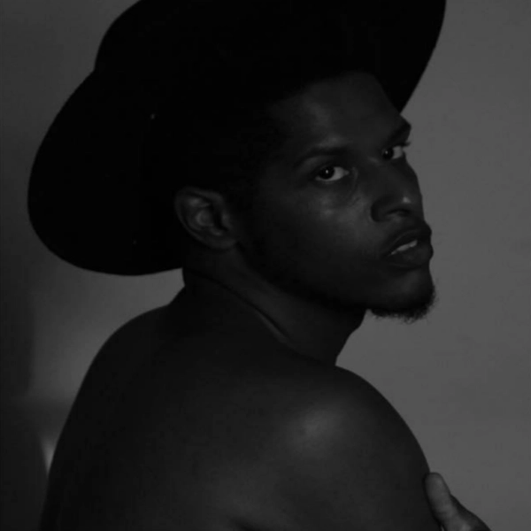 Dim black and white headshot of Malcom-x Betts. They are shirtless, their back facing the camera, their head turned looking into the lens. They are wearing a black brimmed hat.