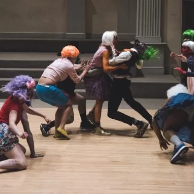 A group of performers wearing multicolored wigs perform a dynamic dance performance on a wooden floor. Two figures are grasping each other in the center of the image, while the other performers crawl and lunge towards them from the wings.