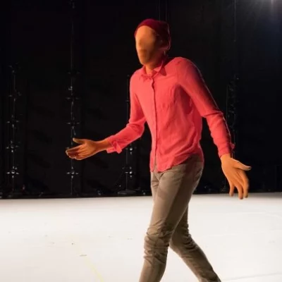 A figure wearing a full coverage body suit, pink button up shirt, and trousers walks across a white stage with outstretched arms.