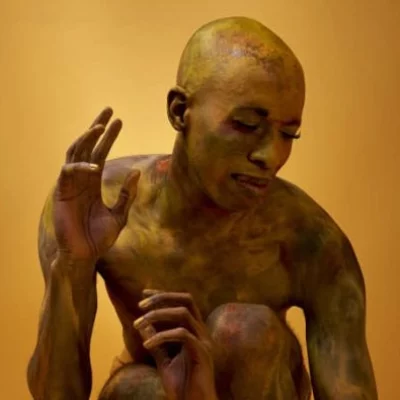 Portrait of Raja Feather Kelly covered in gold body paint, posing squatting down almost nude against a gold background.
