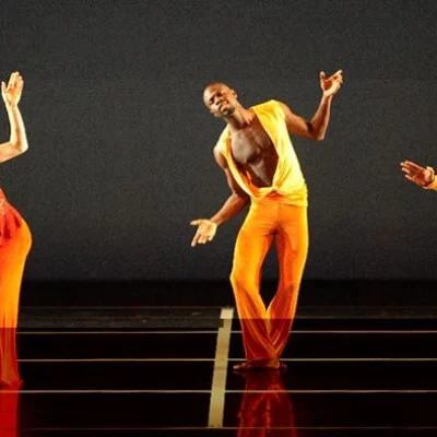 Three dancers in orange costumes tilt their bodies on a black stage
