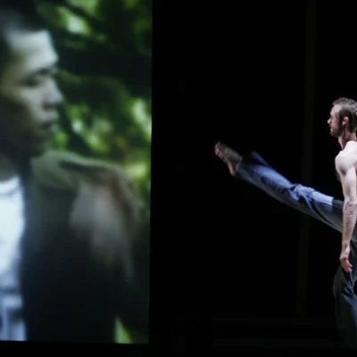 A dancer one a stage extends their leg towards a video projection of a man seeming looking back at them