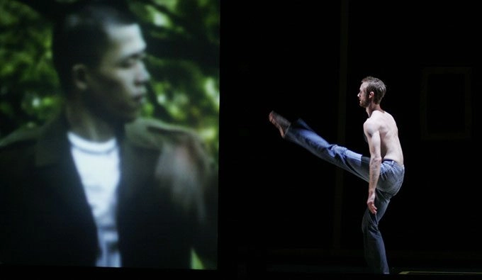 A dancer one a stage extends their leg towards a video projection of a man seeming looking back at them