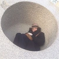 Photo of Bjorn G Amelan sitting in a circle cutout in a concrete wall. They are curled up, wearing a hat and black trench coat.