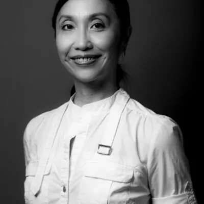 Black and white portrait photo of Janet Wong. They are smiling at the camera with their arms behind their back, wearing a white collarless button up shirt.