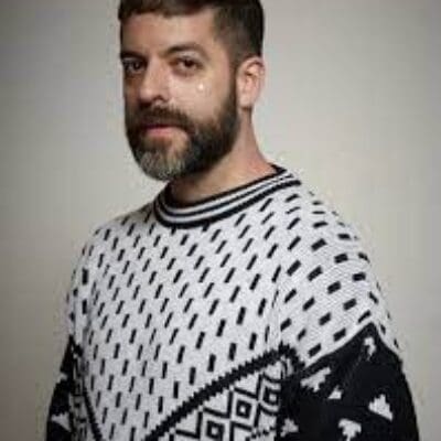 Color portrait photo of Miguel Gutierrez. They are facing the camera, wearing a black and white sweater with a geometric knit pattern against an off-white background.