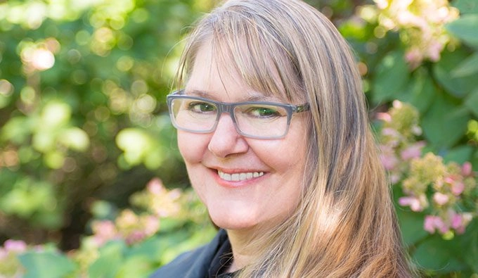 Color portrait photo of RoseAnne Spradlin. They are standing against a green floral background, wearing grey glasses and a black shirt.