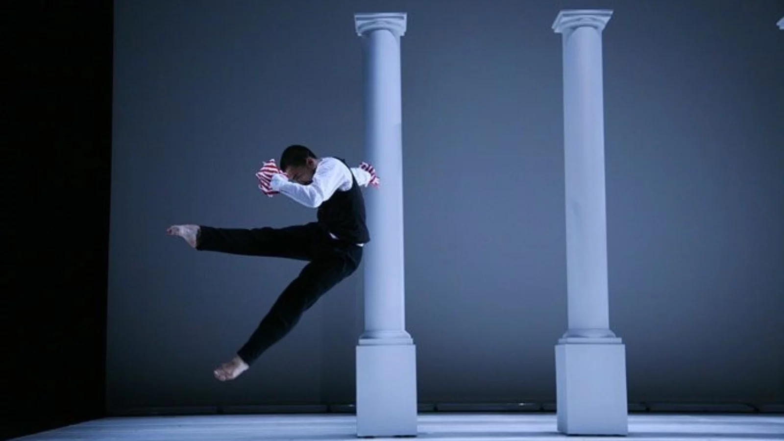 A dancer jumps and kicks their leg in front of a row of white pillars