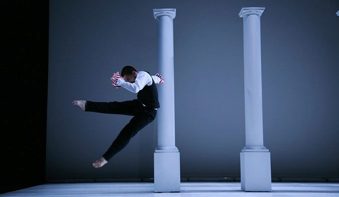 A dancer jumps and kicks their leg in front of a row of white pillars