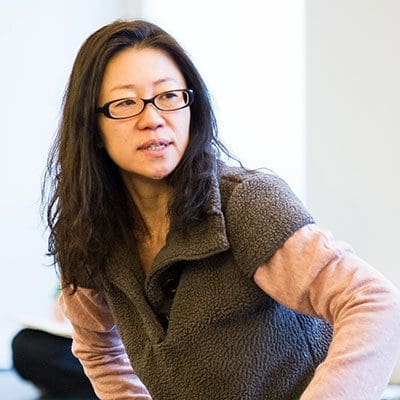 Medium shot of Yasuko Yokoshi. They are sitting in a dance studio, speaking to someone off behind the camera. They are wearing black glasses and a grey sweater.