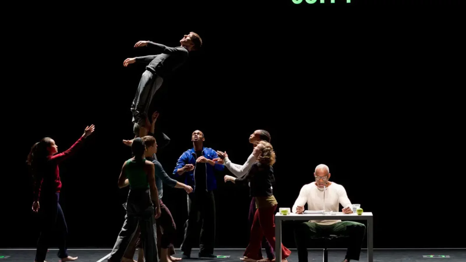 Bill T Jones sits at a desk on stage while dancers perform acrobatic positions behind him