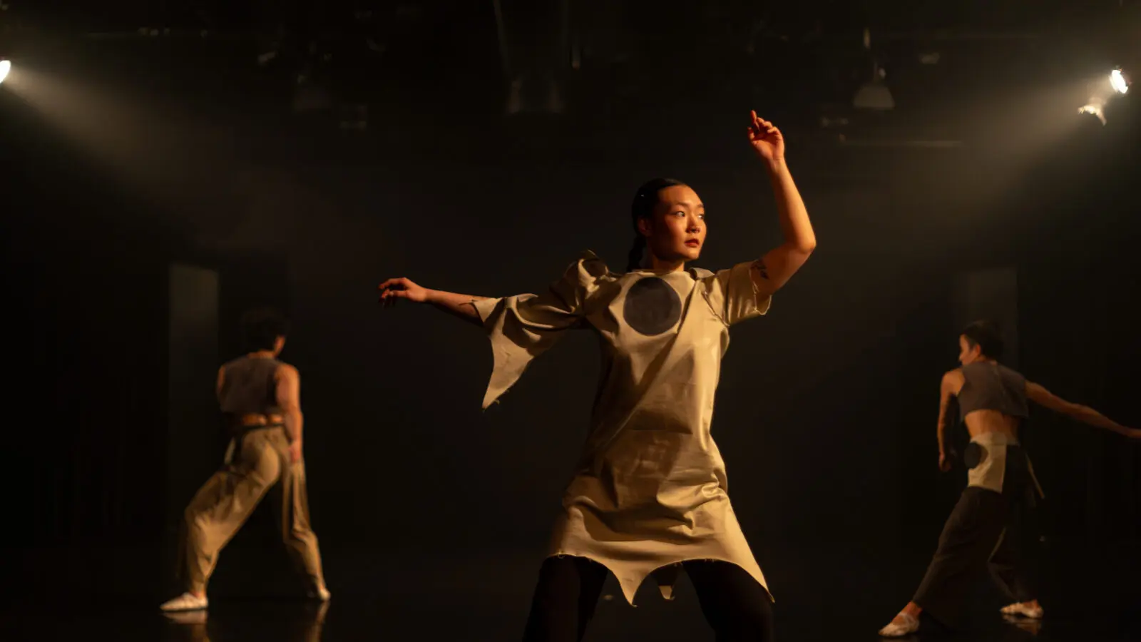 one dancer in the foreground wearing a dress with a circle on the chest, looks to the side with her arms up. two dancers in the background lunge.