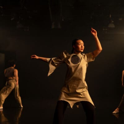 one dancer in the foreground wearing a dress with a circle on the chest, looks to the side with her arms up. two dancers in the background lunge.