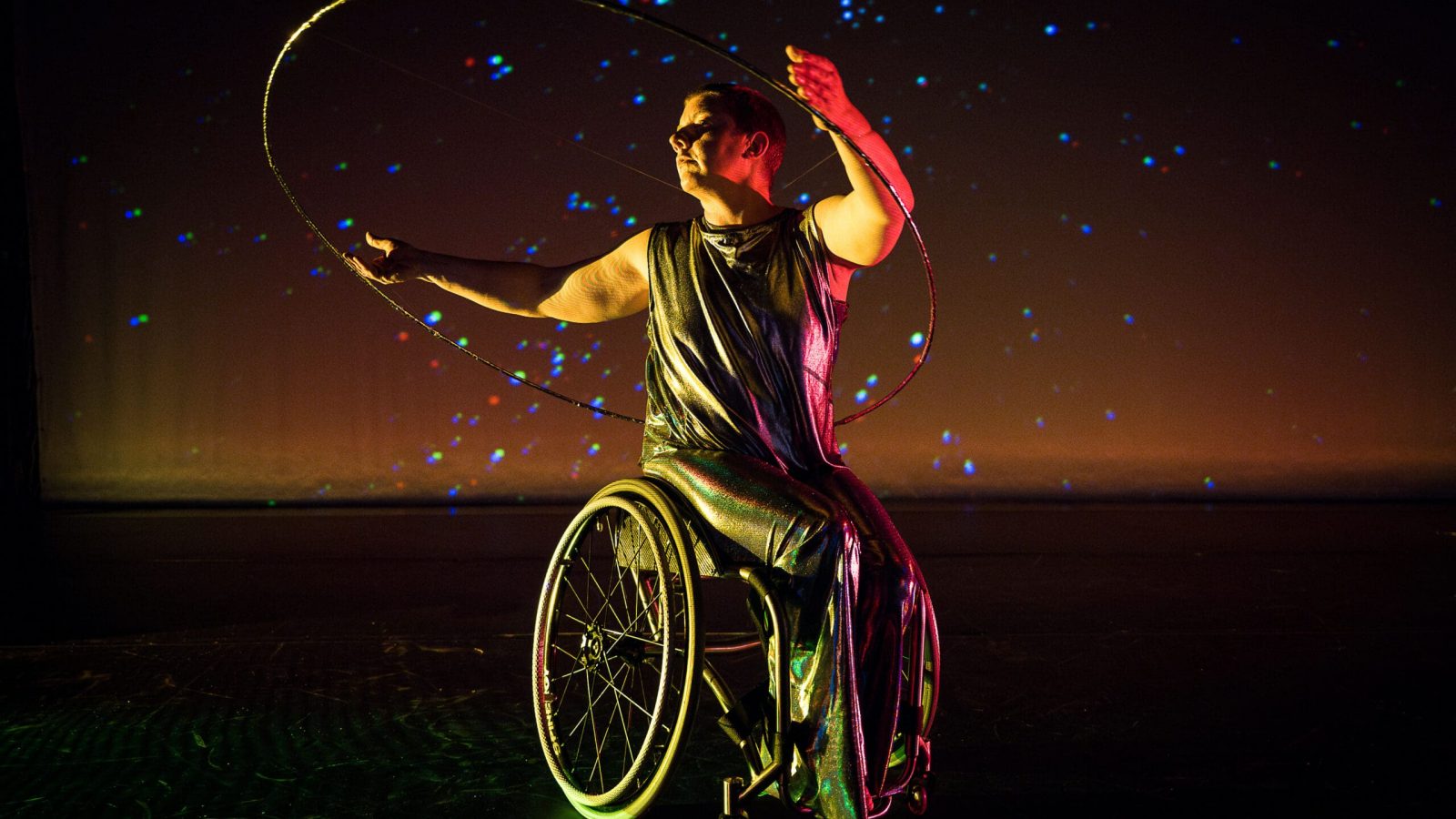 Photo by Cherylynn Tsushima. (Image Description: Laurel Lawson twists their torso as they lift open, curved arms and a large silver hoop.  Laurel is a white person with very short hair; they wear a shimmery sleeveless bodysuit. Projected stardust shines behind them and light reflects off their wheels.)