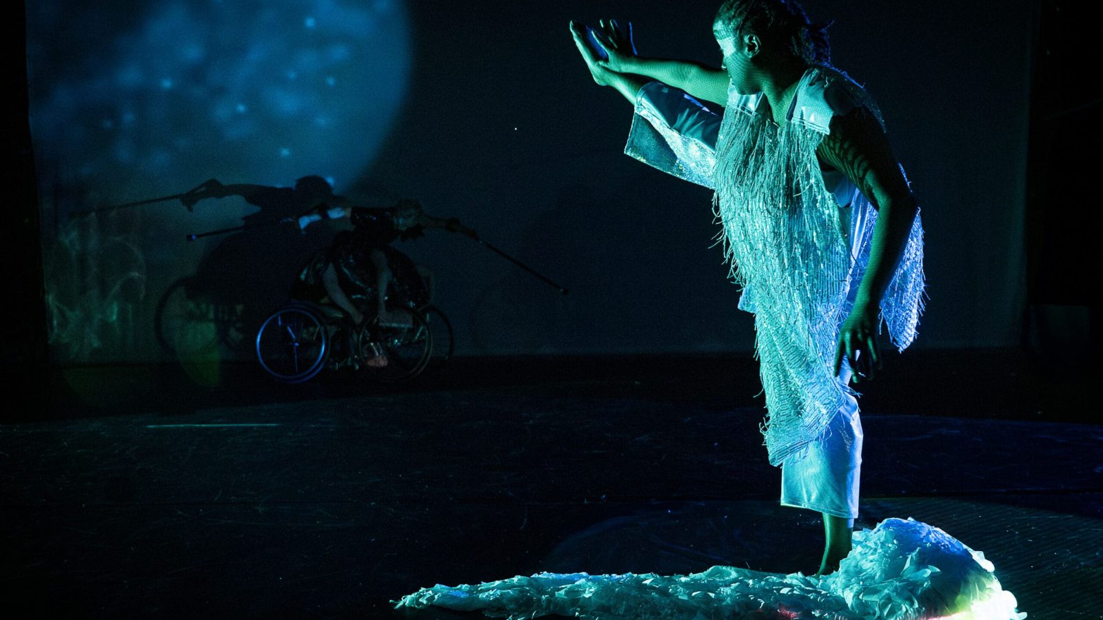 Photo by Cherylynn Tsushima. (Image Description: Kayla Hamilton stands on one leg in a reflective pool; she balances as she lifts her opposite leg high in the air and reaches one hand toward her flat foot. Kayla is a thick, juicy-bodied Black woman, her locs are tied in a short ponytail; she wears a sleeveless white costume of delicate fringe, with white pants. Behind her, a full moon and three figures in the shadows — Laurel Lawson and Tatiana Cholewa are hidden by darkness as they guide their wheels, as Alice Sheppard is perched atop, extending her crutches.)