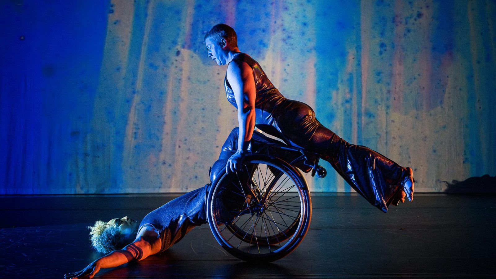 Photo by Cherylynn Tsushima. (Image Description: Laurel Lawson holds their body in a diagonal line as they balance atop Alice Sheppard’s wheels and shins. Alice supports from below as they lay back, arms and torso on the ground, hips  lifted. Alice is a multiracial Black woman with short curly hair, Laurel is a white person with very short cropped hair; they both wear shimmery sleeveless bodysuits. Behind them, a projection of soft blues, purples, and greens looks like rain on a windowpane.)